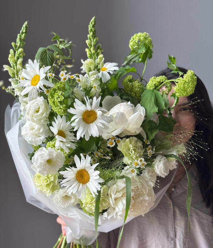 A person holding a large, lush bouquet of white and green flowers, including daisies, roses, and hydrangeas, wrapped in white tissue paper against a plain gray background.